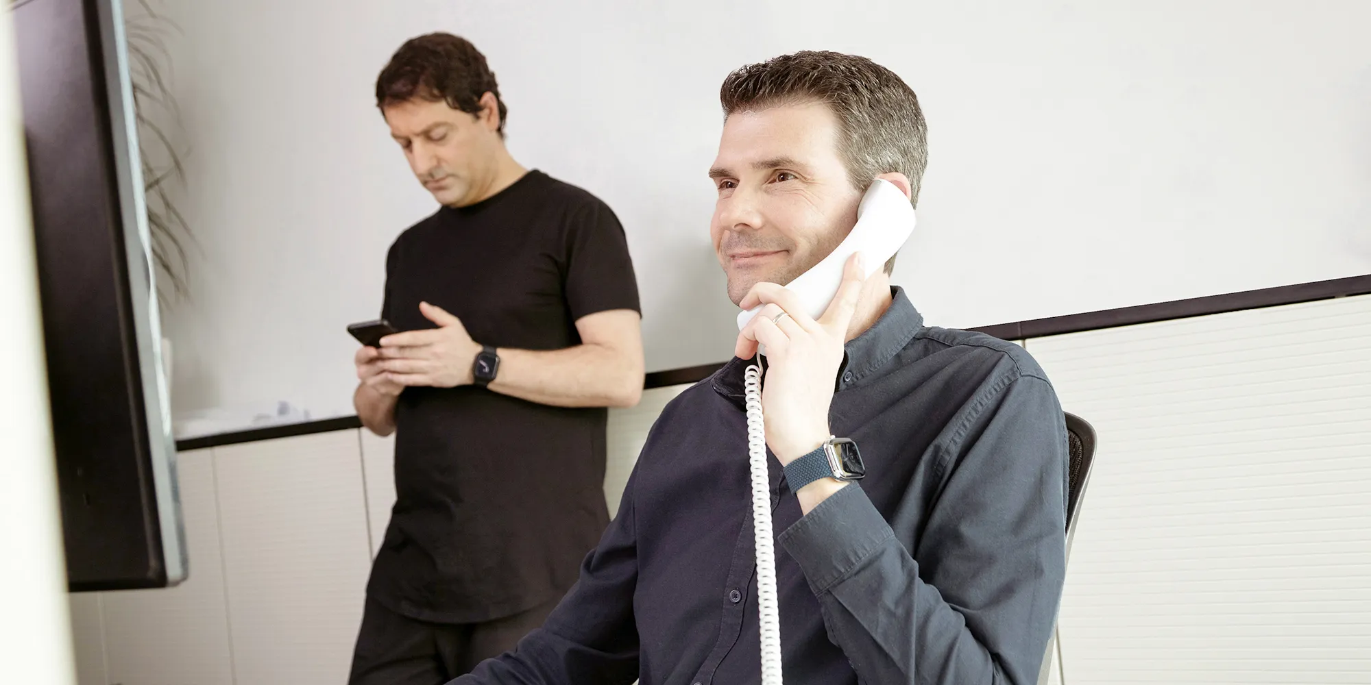 Mann telefoniert mit Tischapparat während ein zweiter am Handy scrollt