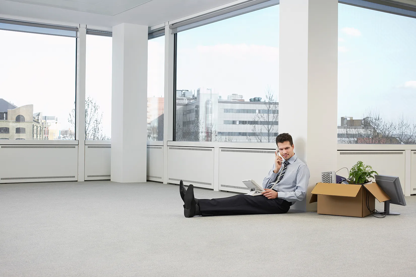 Geschäftsmann sitzt in leerem Büro auf dem Boden und telefoniert mit Festnetzapparat