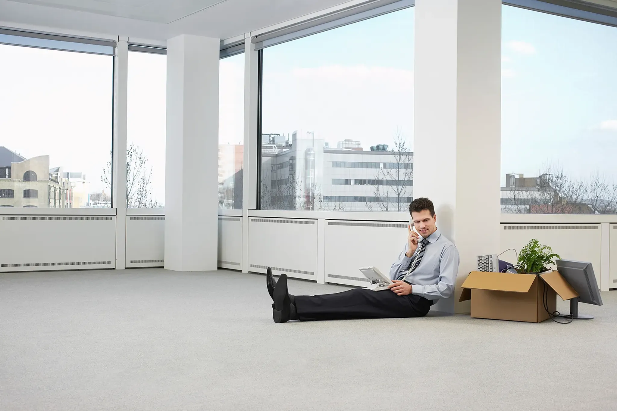 Geschäftsmann sitzt in leerem Büro auf dem Boden und telefoniert mit Festnetzapparat