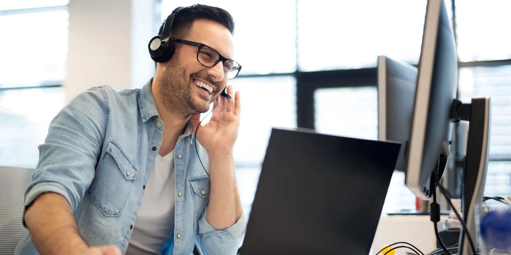 Mann mit Headset schaut auf Laptop und Bildschirm