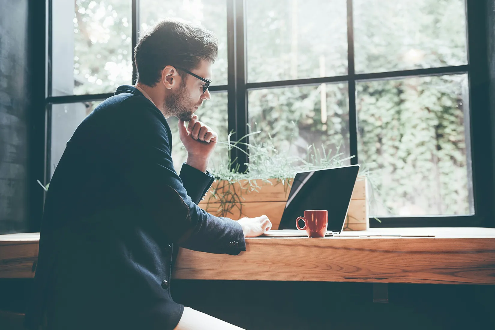Geschäftsmann arbeitet an Fenster mit Laptop und trinkt eine Tasse Kaffee