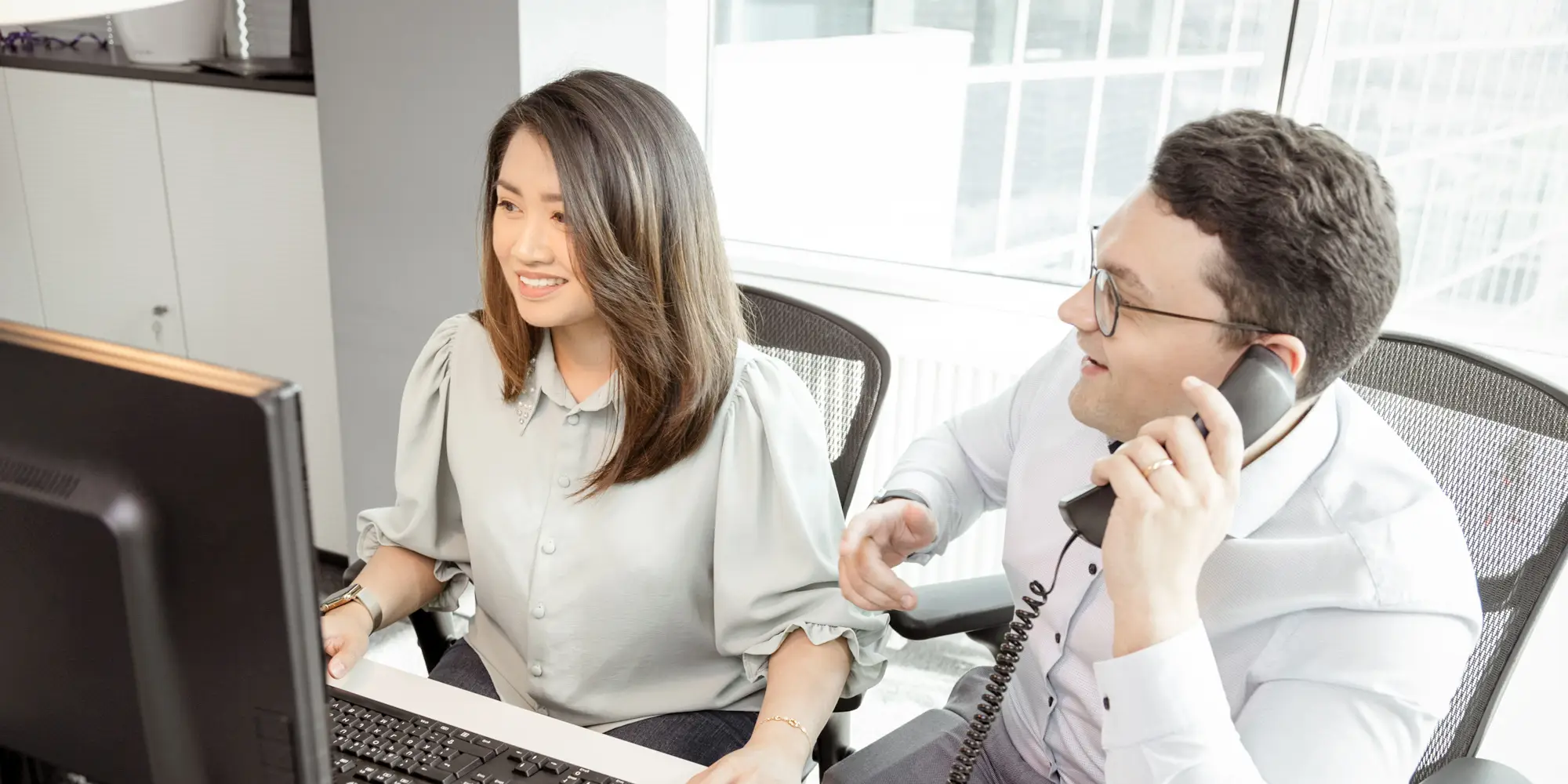 Frau arbeitet im Büro vor Computer während Mann mit Tischapparat telefoniert